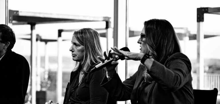 Two women sit beside each other indoors during a panel discussion; one woman listens attentively while the other gestures and speaks into a microphone. The background shows large windows and an industrial view.